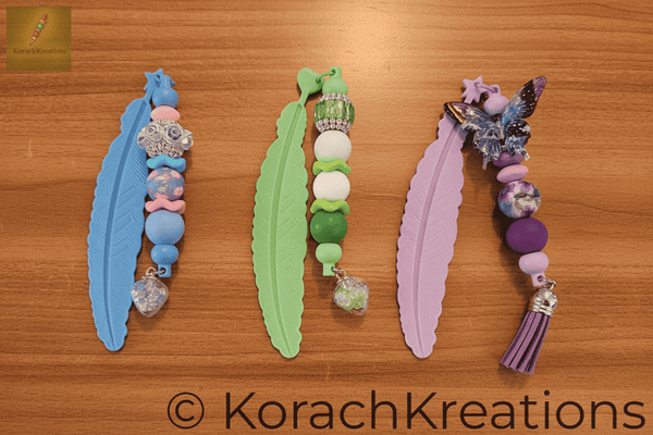 Overhead flat lay of 3 beaded bookmarks arranged side by side on a warm wooden library counter, all beads and tassel designs clearly visible
